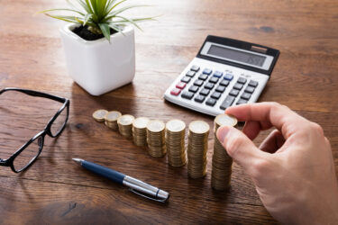Person Hand Calculating On Wooden Desk In Front Of Stacked Coins
