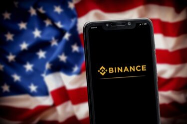 "Binance" logo on the smartphone screen with the national flag of USA at the blurred background.